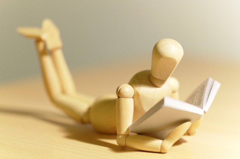 Photo of a wooden model laying belly-down, legs crossed behind them while reading a book