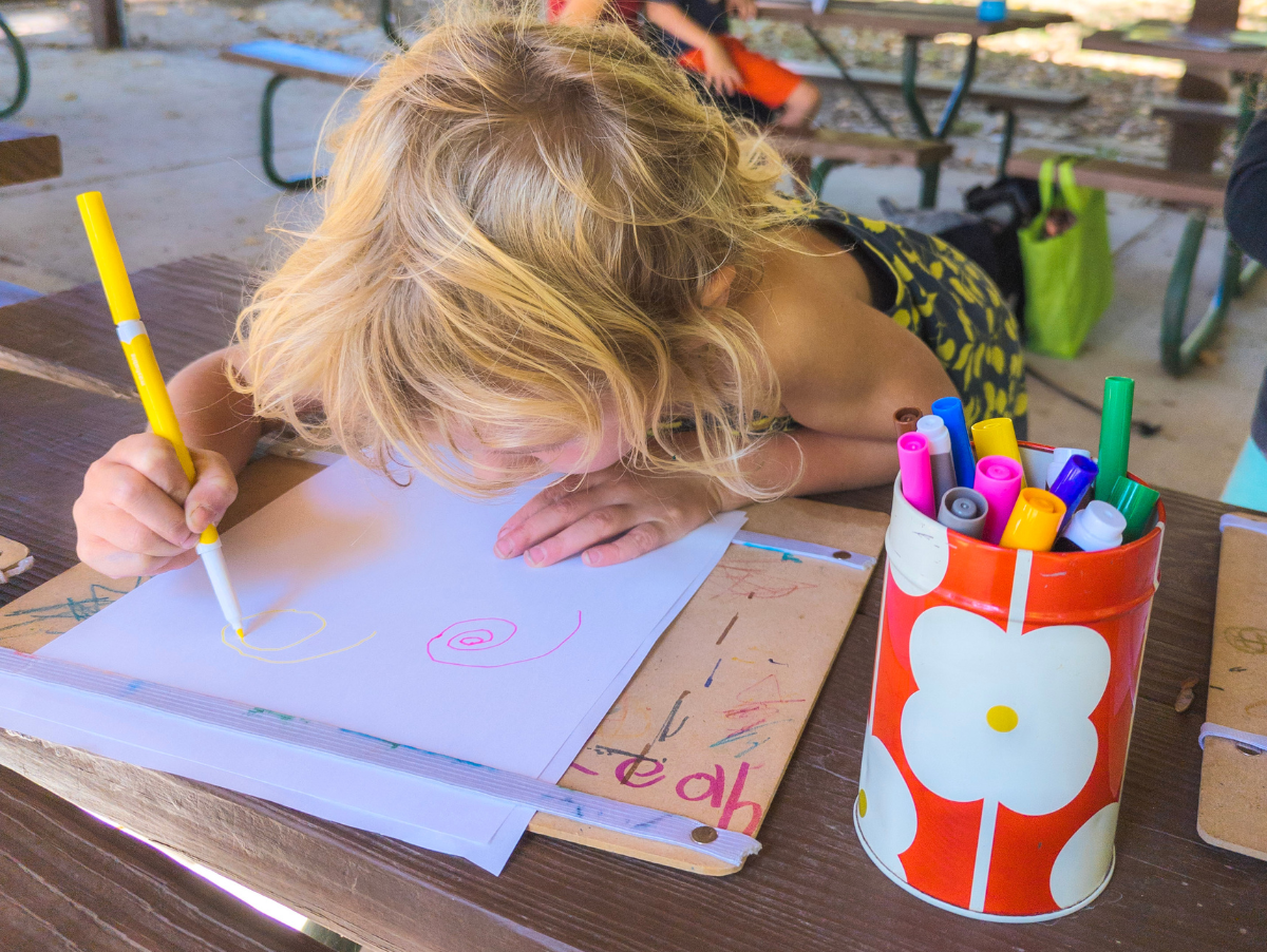 Blonde-haired child drawing spirals on a paper at a picnic table
