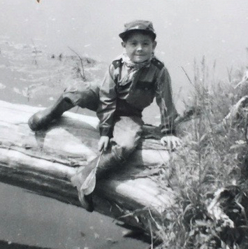 Black and white photo of the author as a young boy looking at the camera and sitting on a log with a pond in the background